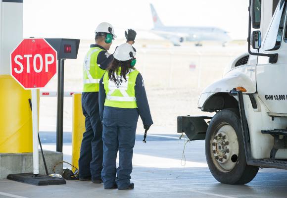 A screening officer is gesturing a vehicle forwards at vehicle screening while another screening officer waits holding an explosives trace detection swab.