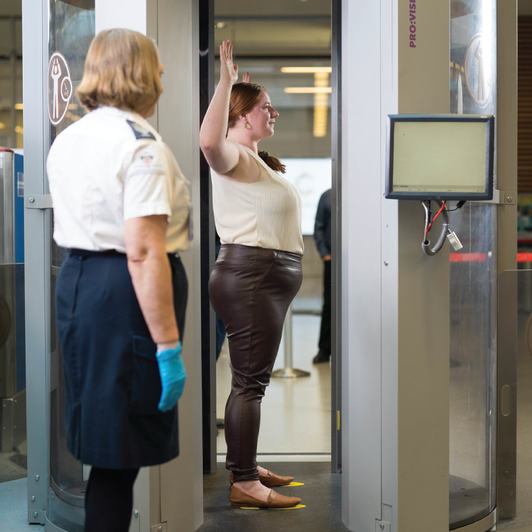 Une passagère se tient debout dans un scanner corporel, les bras levés, sous le regard de deux agents de contrôle.